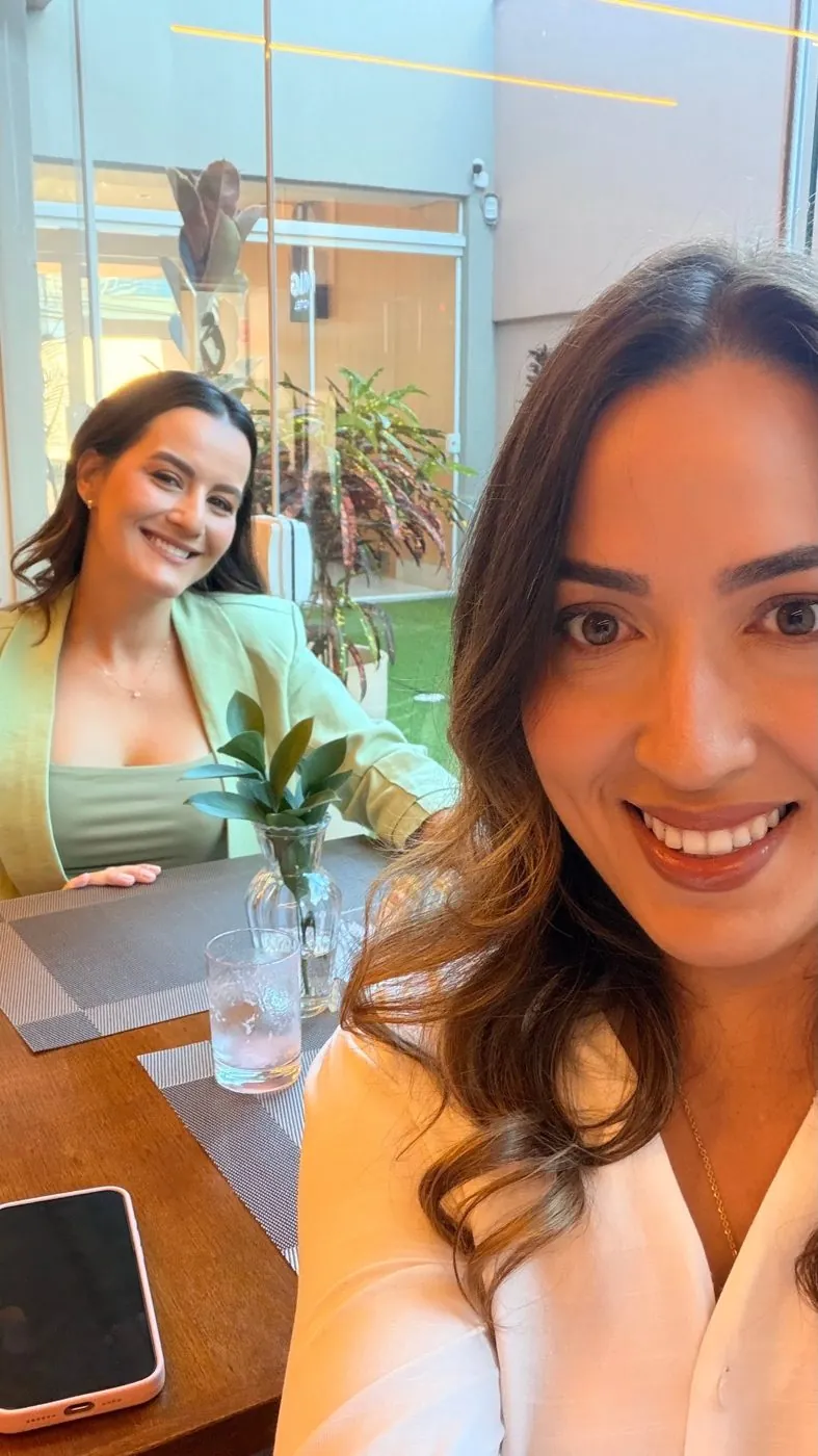 Selfie de Bruna Câmara, em primeiro plano, com Fernanda Tavares sorrindo ao fundo, sentadas à mesa